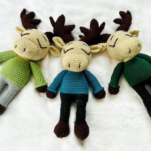 Può includere: Tre bambole di alce all'uncinetto fatte a mano. Ogni bambola ha una testa beige, corna marroni e occhi chiusi. Una bambola indossa una camicia verde e pantaloni grigi, un'altra una camicia blu e pantaloni neri, e la terza una camicia verde e pantaloni neri.