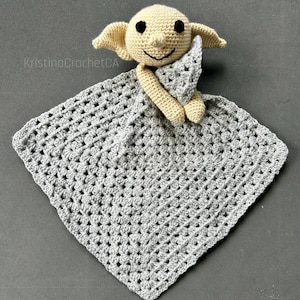 Puede incluir: Una manta de seguridad de crochet hecha a mano con una muñeca beige claro con orejas grandes y una cara sonriente, sosteniendo un triángulo gris. La muñeca está unida a una manta cuadrada de crochet gris. El texto "KristinaCrochetCA" es visible.