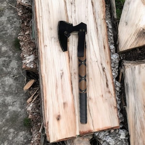 May include: A black axe with a leather handle wrapped in brown leather with a braided design. The axe is resting on a piece of wood.