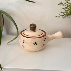 May include: A cream-coloured ceramic pot with a single handle and a matching lid. The pot features a pattern of small dark blue floral motifs and a dark brown rim. A round, dark wood-toned knob sits atop the centre of the lid.