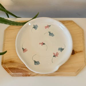 May include: A round, white ceramic spoon rest featuring a scattered pattern of small, delicate pink and light blue flowers with thin green stems painted across the surface. The dish has a smooth, glossy finish and a slightly raised, curved edge.