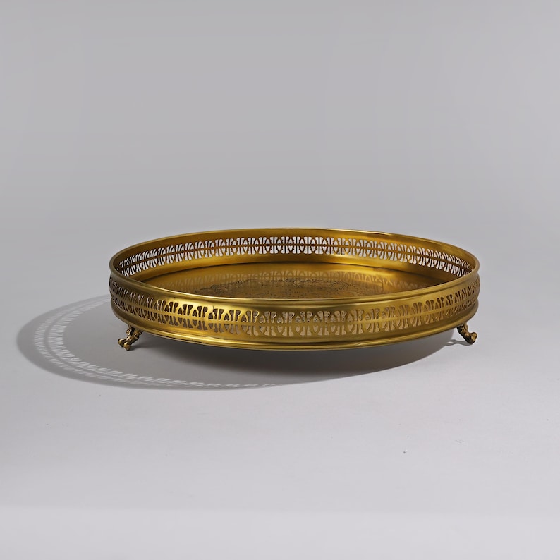 May include: A gold-toned metal serving tray with a decorative, openwork rim and three feet. The tray has a round shape and a smooth surface.