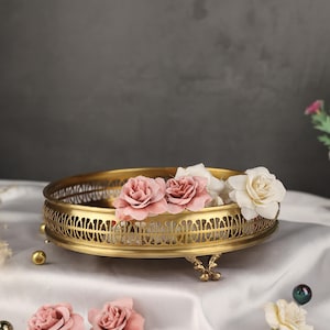May include: A gold metal decorative tray with an intricate design and three legs. The tray is filled with pink and white flowers.