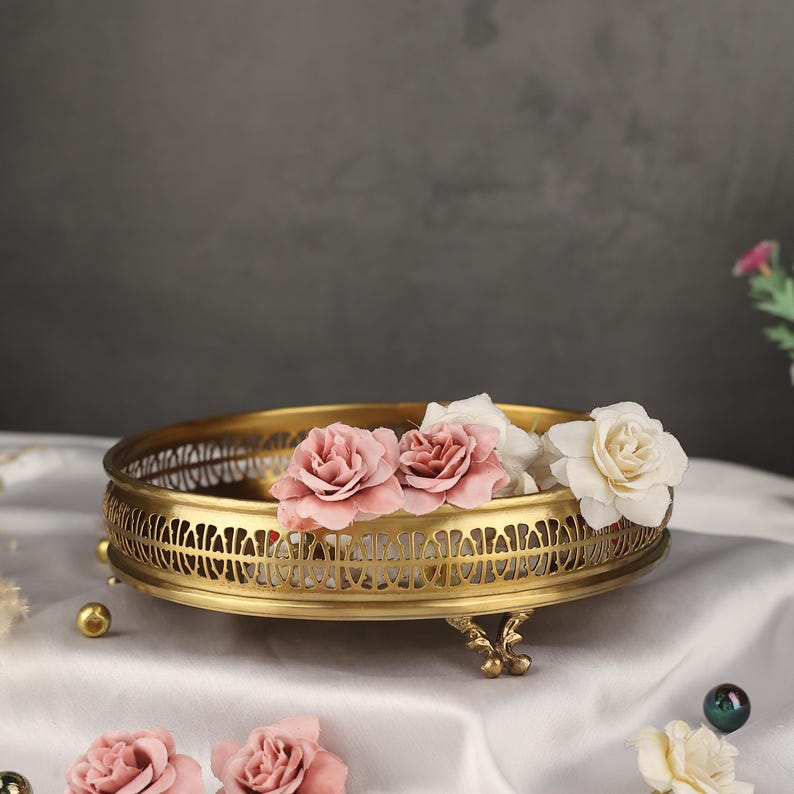 May include: A gold metal decorative tray with an intricate design and three legs. The tray is filled with pink and white flowers.