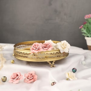May include: A gold metal decorative tray with an intricate design and three legs. The tray is filled with pink and white fabric flowers.