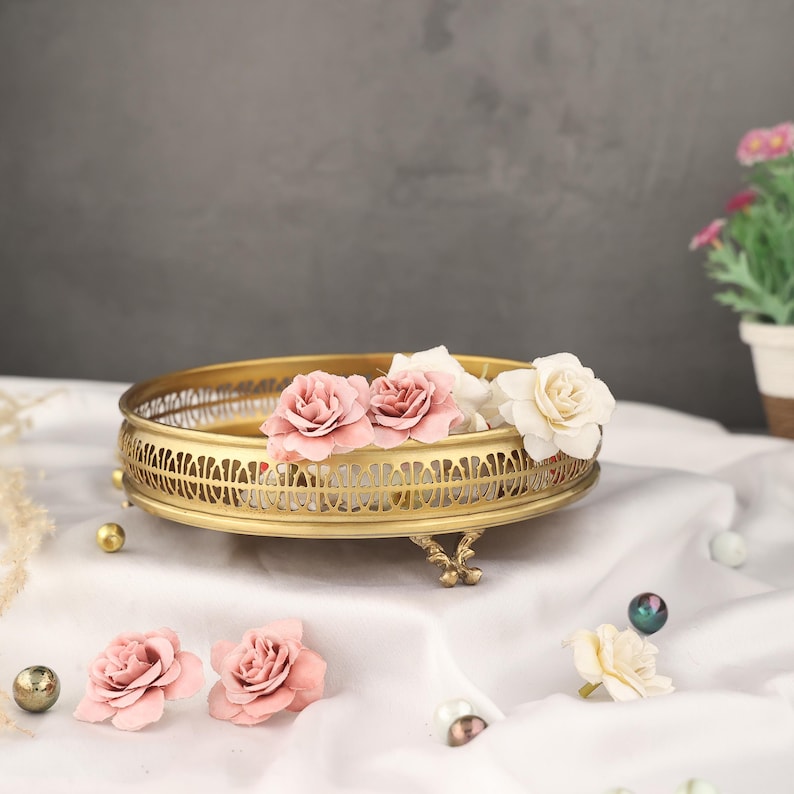 May include: A gold metal decorative tray with an intricate design and three legs. The tray is filled with pink and white fabric flowers.