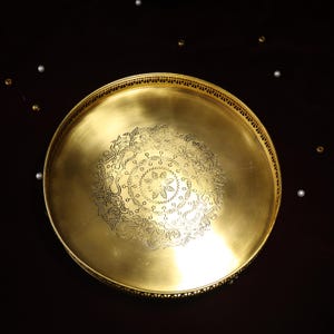 May include: A round, gold-colored metal serving tray with an intricate, engraved design in the center. The tray has a raised rim and a decorative edge.