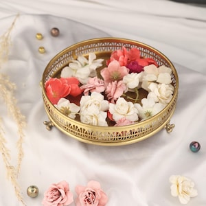 May include: A gold metal decorative tray with a round top and three legs. The tray is decorated with a delicate floral pattern and features a raised rim. The tray is filled with pink and white flowers.