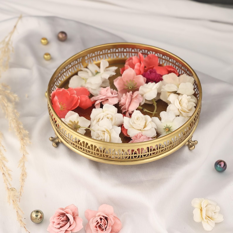 May include: A gold metal decorative tray with a round top and three legs. The tray is decorated with a delicate floral pattern and features a raised rim. The tray is filled with pink and white flowers.