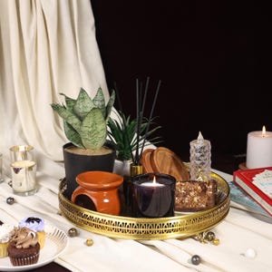 May include: A gold tray with a decorative design holds a variety of items, including a brown and orange ceramic incense burner, a black and clear glass candle, a wooden coaster, a clear glass candle holder, a potted succulent, and a potted snake plant. The tray is on a white surface.