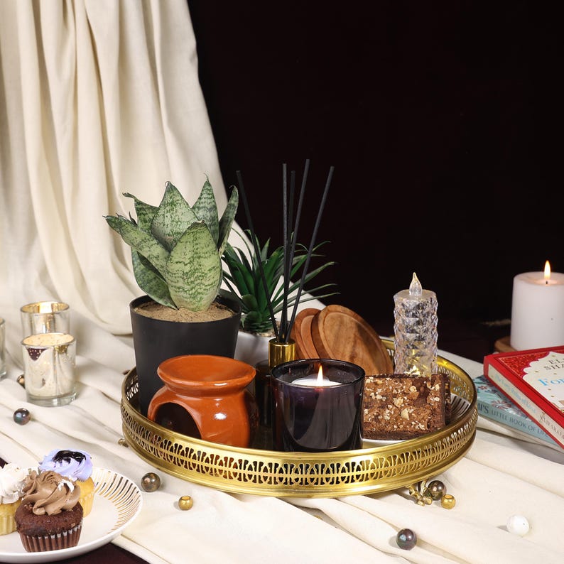 May include: A gold tray with a decorative design holds a variety of items, including a brown and orange ceramic incense burner, a black and clear glass candle, a wooden coaster, a clear glass candle holder, a potted succulent, and a potted snake plant. The tray is on a white surface.