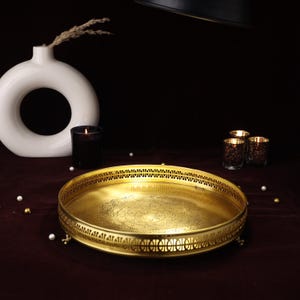 May include: A gold-toned metal serving tray with an intricate design and a raised rim. The tray has a decorative pattern on the bottom and sits on four small feet.