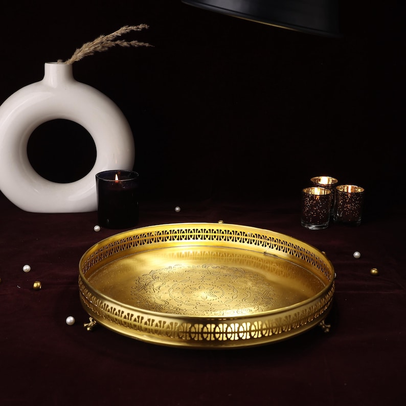 May include: A gold-toned metal serving tray with an intricate design and a raised rim. The tray has a decorative pattern on the bottom and sits on four small feet.