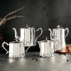 May include: A set of four silver-colored teapots and a creamer with ornate handles and lids. The teapots have a geometric design and are arranged on a wooden surface. The background is a dark, textured wall.