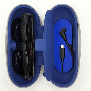 Könnte beinhalten: Geöffnete, graue Reißverschlusstasche mit blauem Innenraum, die eine schwarze Smart-Brille und ein schwarzes USB-C-Ladekabel enthält. Die Brille hat dunkle Gläser und ein schlankes, modernes Design. Die Tasche ist oval.