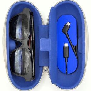 以下が含まれることがあります： 青い内装のグレーのジッパー付きケースが開いています。中には黒いフレームのメガネと黒い充電ケーブルが入っています。ケースは楕円形で、青い仕切りがあります。メガネは濃い色のレンズです。