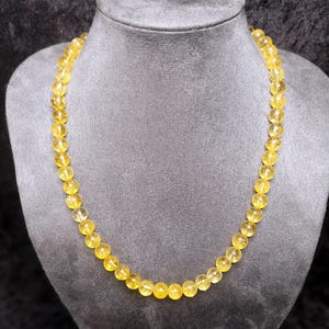 Könnte beinhalten: Eine Halskette aus zahlreichen runden, gelben Perlen. Die Perlen sind zu einer kompletten Halskette aufgereiht. Die Halskette wird auf einer grauen Mannequin-Büste präsentiert, die das Design und die Farbe des Schmucks hervorhebt.