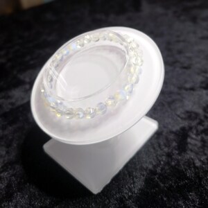 May include: A clear, iridescent beaded bracelet displayed on a white, angled jewelry stand. The bracelet features small, round beads with a subtle, shimmering effect. The stand is a simple, modern design.