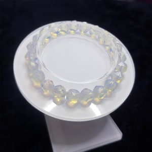 May include: Two opalescent beaded bracelets are displayed on a white, tiered stand. The beads have a faceted, irregular shape and a soft, translucent quality with hints of yellow and white. The stand is set against a dark background.