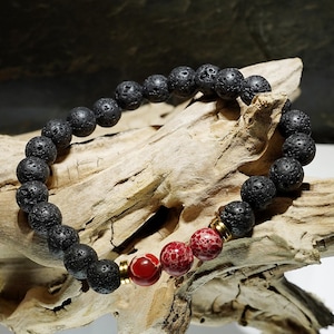 Könnte beinhalten: Ein Armband aus schwarzen Lavasteinen mit einem roten und weißen Achat-Perlen. Das Armband liegt auf einer Holzoberfläche.