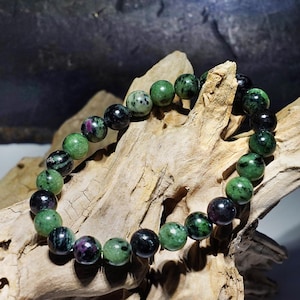 Op de afbeelding: Een armband met ronde edelsteen kralen, voornamelijk groen met zwarte en paarse insluitsels. De kralen zijn gerangschikt in een cirkel. De armband wordt gepresenteerd op een stuk drijfhout, dat de natuurlijke kleuren benadrukt.