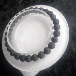 Könnte beinhalten: Ein Armband aus schwarzen Lavasteinperlen, das auf einem klaren, kreisförmigen Ständer präsentiert wird. Das Armband besteht aus runden, dunkelgrauen Perlen. Der Ständer hat einen gewellten Rand und ein weißes, mattiertes Aussehen.