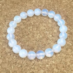 May include: A white opalite bead bracelet. The round beads are strung together, forming a circular shape. The beads have a milky, iridescent quality, reflecting light. The bracelet is displayed on a textured, brown surface.
