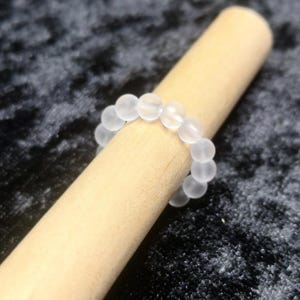 Op de afbeelding: Een witte, gematteerde glazen kralenring. De ring is gemaakt van kleine, ronde kralen die aan elkaar geregen zijn om een cirkel te vormen.