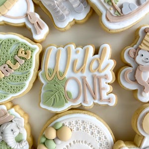 Puede incluir: Galletas de azúcar decoradas con temática de jungla. Las galletas incluyen una cebra, un mono, un elefante y las palabras "Wild One" con un diseño de hoja. Otras galletas tienen las palabras "Happy Birthday Charlie". Las galletas están decoradas con glaseado blanco, marrón claro, verde y melocotón.