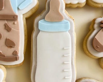 Op de afbeelding: Een witte babyfles-koekje versierd met bruine en blauwe glazuur. De fles heeft een bruine speen en een blauwe ring rond de bovenkant. Het koekje is lichtbruin van kleur en heeft een licht ruwe textuur.