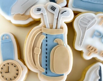 Puede incluir: Una colección de galletas de azúcar decoradas con temática de golf. Las galletas incluyen una bolsa de golf con palos, un reloj de pulsera y otras formas relacionadas con el golf. Las galletas están decoradas con glaseado azul claro, beige y blanco.