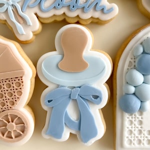 Può includere: Primo piano di biscotti di zucchero decorati a tema baby shower. I biscotti includono un ciuccio, una carrozzina e la parola "bloom" in glassa blu. I biscotti sono decorati con glassa azzurra e beige.