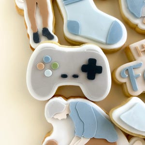 以下が含まれることがあります： デコレーションされた砂糖クッキーのクローズアップ。クッキーは、ビデオゲームコントローラー、靴下、人の形をしています。コントローラーはグレーで、カラフルなボタンが付いています。靴下は水色です。人は青いシャツと茶色のズボンを着ています。