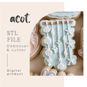 May include: A decorated sugar biscuit with a light blue background, featuring white seashell and starfish designs. The biscuit is shaped like a rectangle with a scalloped edge. The image also includes text that says "acot."