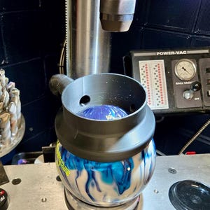 May include: A blue and white bowling ball is being drilled with a drill press. The ball is held in place by a black plastic funnel with a hose attached to it. The drill press has a control panel labeled "POWER-VAC" with a gauge and a switch labeled "BALL VAC" and "OFF".