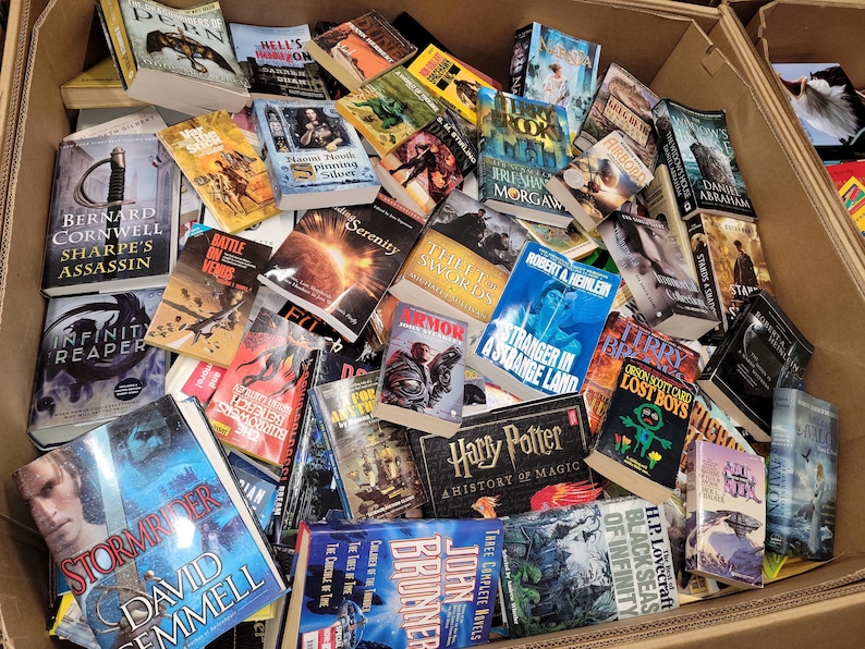 Pode incluir: Uma caixa de cart&atilde;o cheia de uma variedade de livros de bolso, incluindo fantasia, fic&ccedil;&atilde;o cient&iacute;fica e fic&ccedil;&atilde;o hist&oacute;rica. Os t&iacute;tulos vis&iacute;veis incluem "Sharpe's Assassin", "Infinity Reaper", "Battle in Venus", "Serenity", "Stranger in a Strange Land", "Harry Potter", "Black Seas of Infinity" e "Stormbringer".