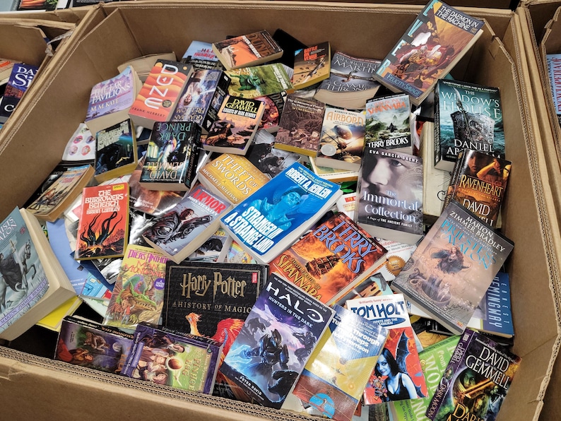 Pode incluir: Uma caixa de cart&atilde;o cheia de uma variedade de livros de bolso. Os livros s&atilde;o principalmente de fantasia e fic&ccedil;&atilde;o cient&iacute;fica, com t&iacute;tulos como "Stranger in a Strange Land", "The Chronicles of Narnia", "Harry Potter", "Dune", "The Demonata", "Ravenheart", "Immortals", "Halo" e "Shannara".