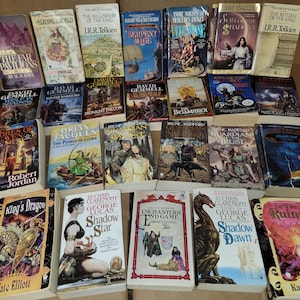 May include: A collection of fantasy and science fiction paperback books with various covers featuring dragons, wizards, and other mythical creatures. The books are arranged in a grid pattern on a brown surface.