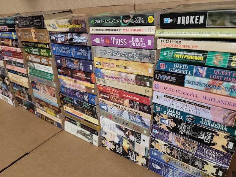 May include: A large collection of paperback books stacked on a shelf. The books are mostly fantasy and science fiction novels, with titles like "The Hero of Ages", "Star of Danger", "The Last Guardian", and "Waylander".