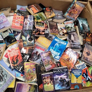 Pode incluir: Uma caixa transbordando com uma variedade de livros de bolso, muitos com temas de fantasia e fic&ccedil;&atilde;o cient&iacute;fica. Os t&iacute;tulos incluem "Dune", "The Far Pavilions", "Prince of Darkness", "Armor", "Stranger in a Strange Land", "Immortals Collection", "Shannara", "Halo: Hunters in the Dark" e "A History of the World".