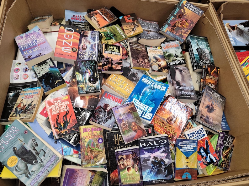 Pode incluir: Uma caixa transbordando com uma variedade de livros de bolso, muitos com temas de fantasia e fic&ccedil;&atilde;o cient&iacute;fica. Os t&iacute;tulos incluem "Dune", "The Far Pavilions", "Prince of Darkness", "Armor", "Stranger in a Strange Land", "Immortals Collection", "Shannara", "Halo: Hunters in the Dark" e "A History of the World".