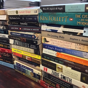 May include: A stack of paperback books with various titles and authors. The books are arranged vertically, with the spines facing outward. Some of the titles include "The Lynching Waltz", "A Single Breath", "The One That Got Away", and "A Prayer for Owen Meany".