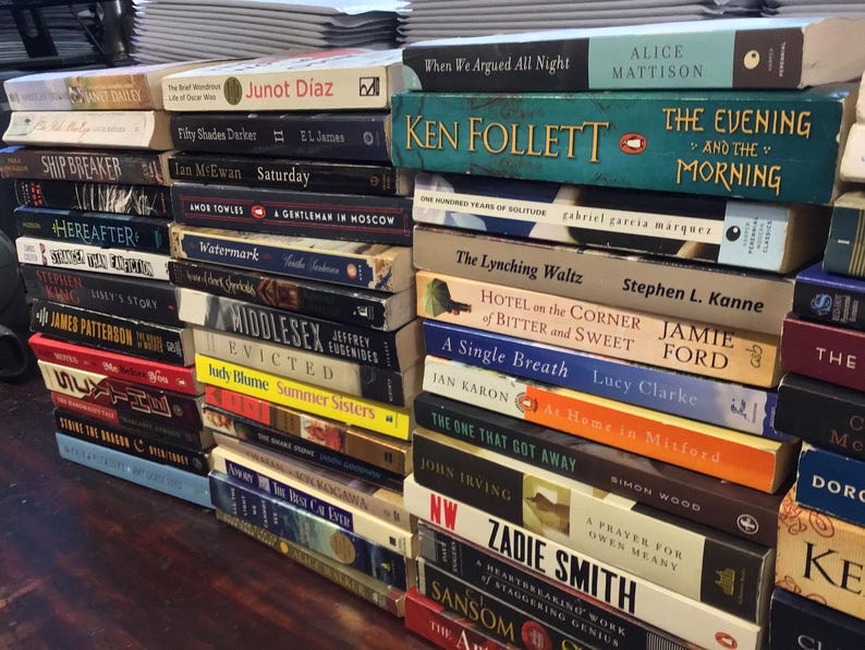 Op de afbeelding: Een stapel paperbacks met verschillende titels en auteurs. De boeken zijn verticaal gerangschikt, met de ruggen naar buiten. Sommige van de titels zijn "The Lynching Waltz", "A Single Breath", "The One That Got Away" en "A Prayer for Owen Meany".