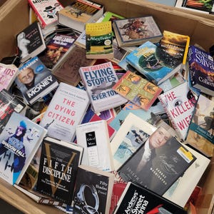 May include: A cardboard box overflowing with a variety of paperback books. The books are stacked on top of each other and some are sticking out of the box. The titles of some of the books include "The Dying Citizen", "The Greatest Flying Stories Ever Told", "The Killer's Wife", and "Fifty Shades Freed".