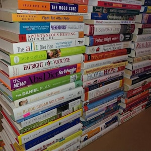 May include: A large stack of books with various titles related to health, diet, and wellness. The spines display titles like "Sonoma Diet," "Eat Right 4 Your Type," and "The Daniel Plan." The books are stacked against a cardboard surface.