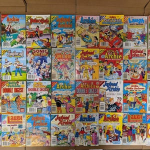 May include: A collection of vintage Archie Comics and related titles, featuring colorful covers with characters like Archie, Betty, Veronica, and Jughead. The comics are arranged on a cardboard surface, showcasing various issues and titles.