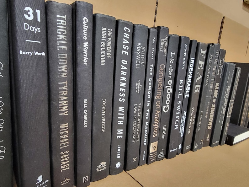 Pu&ograve; includere: Una collezione di libri con copertina rigida di colore grigio scuro e scritte bianche. I titoli includono "31 Days", "Trickle Down Tyranny", "Culture Warrior" e "Chase Darkness With Me". I libri sono disposti su uno scaffale.