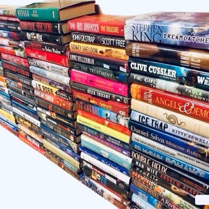 May include: A large stack of paperback books, mostly fiction, with various titles and authors visible. The books are arranged in a vertical stack, with the spines facing outward. The books are a mix of colors, including red, blue, green, and black.