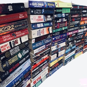 May include: A large stack of paperback books, mostly in shades of blue, black, and red, with some white and yellow spines. The books are arranged in a vertical stack, with the spines facing out. The titles of some of the books are visible, including "The Da Vinci Code", "Medusa", "Hard Evidence", "The Perfect Girlfriend", and "The Throat".