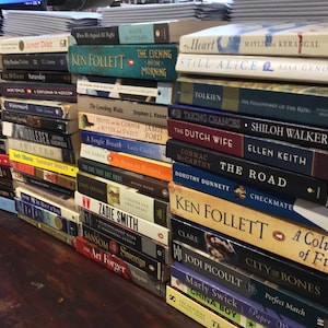 Op de afbeelding: Een stapel paperbacks met verschillende titels en auteurs. De boeken zijn verticaal gestapeld, met de titels naar buiten gericht. Enkele van de titels zijn "The Evening and the Morning", "Still Alice", "The Fellowship of the Ring", "The Road", "Checkmate", "A Column of Fire", "City of Bones" en "China Boy".